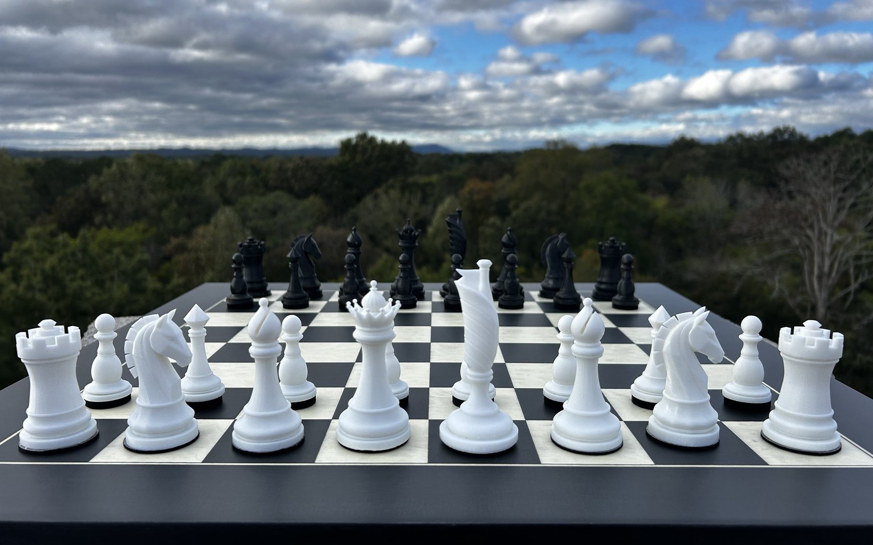 A Flag Chess set in its starting position on a black-and-white board, seen low from the White side, with all four Sentinels visible, the Flag’s pennant in profile at the center, and a forested ridgeline and clouds behind